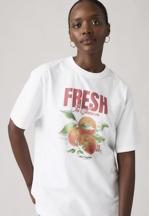 Femme portant un T-shirt blanc avec un texte rouge "FRESH In Season" et un graphique de trois pêches avec des feuilles et des fleurs sur un fond uni.