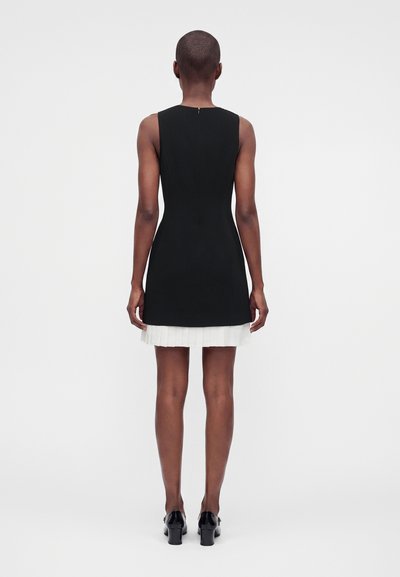 Robe noire sans manches et ajustée avec un ourlet plissé blanc portée par une personne debout vue de dos sur un fond uni.