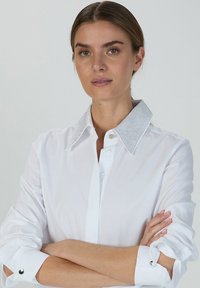 Weißes Hemd mit einem strukturierten, glitzernden Kragen; mit Knopfleiste und aufgerollten Ärmeln mit silbernen Akzenten. Glattes Material.