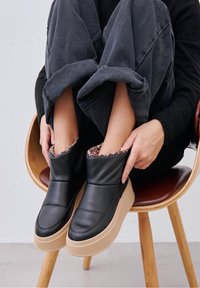 Persona sentada en una silla de madera, vistiendo botas de tobillo acolchadas negras con suelas beige y forro de leopardo, con las piernas dobladas y las manos sosteniendo las botas.