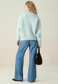 Frau mit lockigem blonden Haar trägt einen hellblauen, flauschigen Pullover, weite blaue Jeans und hält eine dunkelbraune, mit Nieten besetzte Handtasche in der Hand, während sie sich von der Kamera abwendet.