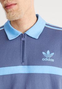 Modré polo tričko na zip s světle modrým límcem, texturovanou látkou, s bílým vodorovným pruhem a vyšitým logem Adidas.