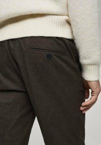 Braun kariertes Hosen mit strukturiertem Stoff, ausgestattet mit einer Seitentasche und einem Knopfdetail, kombiniert mit einem weichen cremefarbenen Pullover.