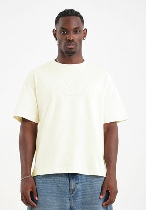 Hombre con el cabello trenzado lleva una camiseta oversize color crema con un sutil logo "AVX" y vaqueros azules sobre un fondo blanco.