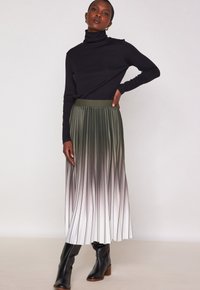 Maglione nero a collo alto con maniche lunghe, abbinato a una gonna plissettata in gradiente che passa dal verde scuro al bianco. Stivali neri con tacco.