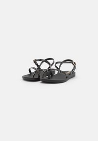 Black flat sandals made of plastic with an open-toe design. Crossed straps and a buckle detail at the ankle. Textured footbed.