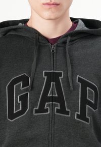 Gros plan sur une personne portant un sweat à capuche zippé gris foncé avec de grandes lettres noires en contours "GAP" sur le devant et un t-shirt bordeaux en dessous.