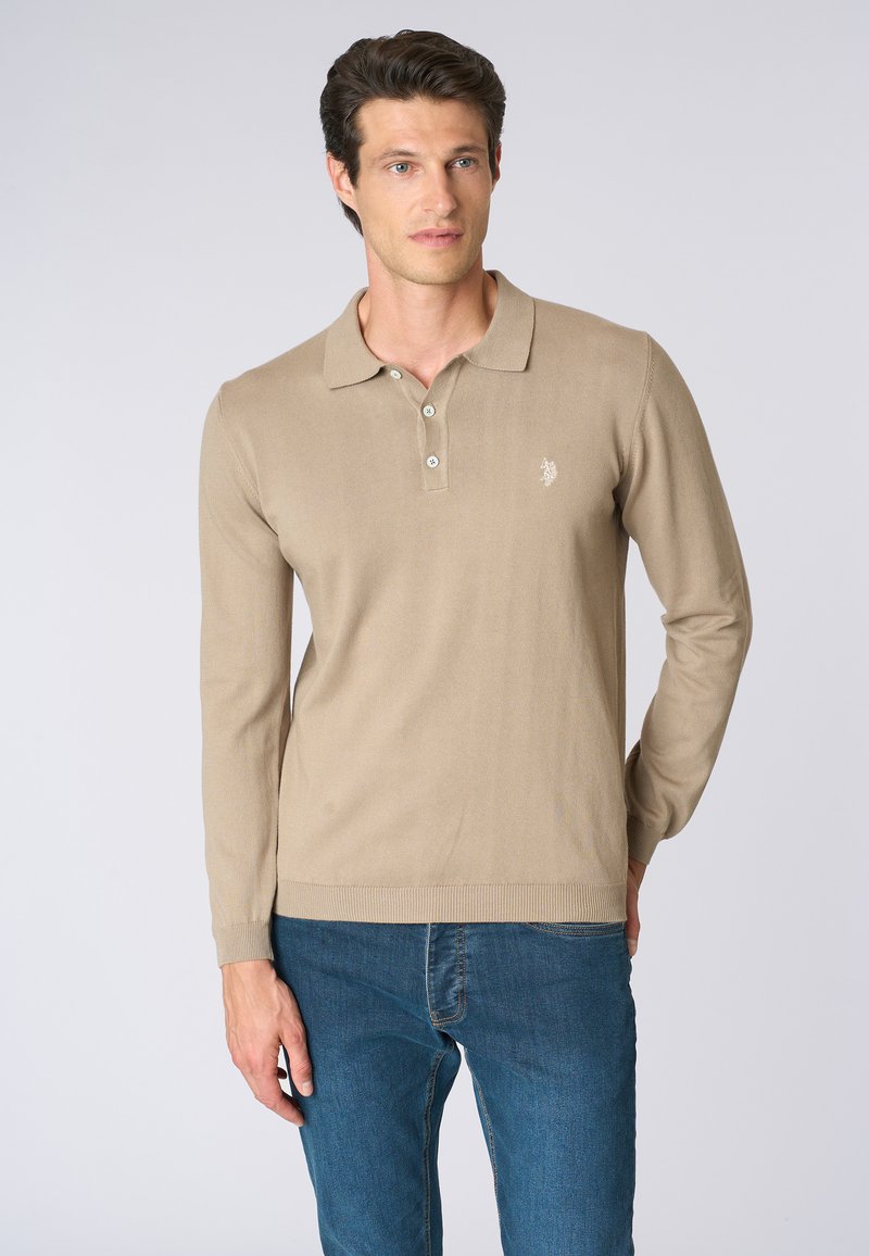 Polo a maniche lunghe beige con texture a coste, apertura con tre bottoni e piccolo logo ricamato sul lato sinistro del petto. Abbinato a jeans blu.