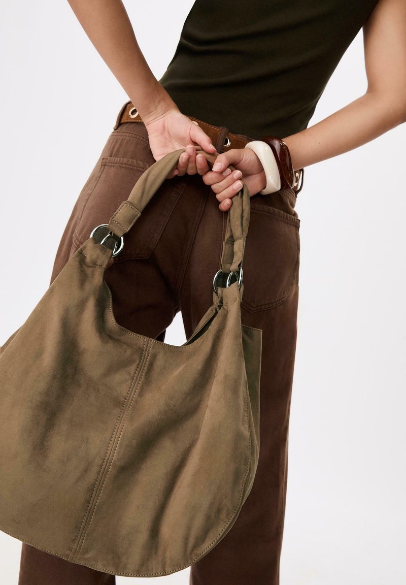 Borsa in suede marrone con una grande forma hobo. Presenta dettagli di cucitura e hardware in tonalità argento sulla tracolla. Tessuto morbido e casual.