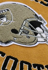 Gray and black embroidered football helmet on orange fabric, featuring bold black lettering and textured details on the helmet design.