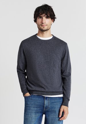 Mann mit dunklem, lockigem Haar, der einen dunkelgrauen Pullover über einem weißen Hemd und blauen Jeans trägt und mit einer Hand in der Tasche steht.