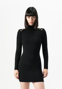 Donna con capelli scuri e lisci che indossa un mini dress a collo alto a coste nero con maniche lunghe e bottoni decorativi dorati sulle spalle.