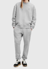 Personne portant un sweat-shirt gris clair et un pantalon de survêtement assorti, les mains dans les poches, assortis de baskets noires et blanches.