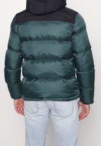 Grön puffjacka med svart huva och axelparti, med en quiltad textur och elastiska manschetter. Kallas med ljusblå jeans.