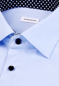 Camicetta azzurro chiaro con una texture liscia, bottone nero e colletto blu navy con un piccolo motivo a pois bianchi. Etichetta del marchio visibile.