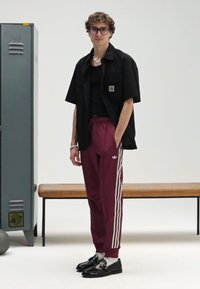 Czarna koszula z krótkim rękawem na czarnym topie, bordowe spodnie dresowe z białymi paskami i czarne buty. W tle znajduje się ławka i szafka.