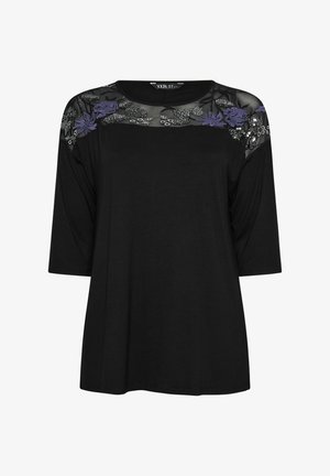 Camiseta negra de manga corta, con un yugo de malla negra transparente adornado con bordados de flores en púrpura y plata. Corte regular.