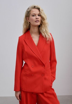 Žena s vlnitými blond vlasmi má na sebe jasne červené dvojradové sako a zodpovedajúce červené nohavice, stojí na jednoduchom svetlosivom pozadí.