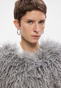 Szary, kudłaty płaszcz z miękką, teksturowaną powierzchnią. Modelka nosi kolczyki w kształcie kół, z neutralnym wyrazem twarzy i krótkimi włosami.