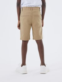 Pantaloni corti khaki con una superficie liscia, dotati di due tasche posteriori e una cintura, abbinati a sneaker bianche. Vista da dietro.