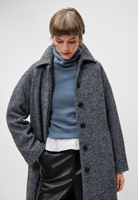 Pelēkā un tumši zilos krāsās ar pūkaini rakstu apmetnis, ar apkakli, priekšējiem pogām un teksturētu audumu, uzvilkts virs zilā augstās kakla un baltās kreklā.