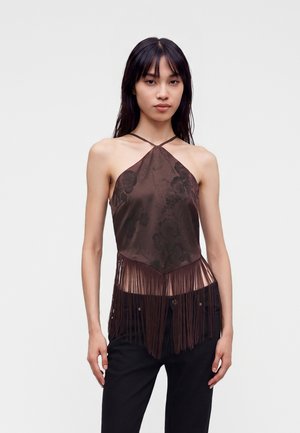 Femme aux longs cheveux foncés portant un haut marron à col licou avec motif floral et détails à franges, associé à un pantalon noir.