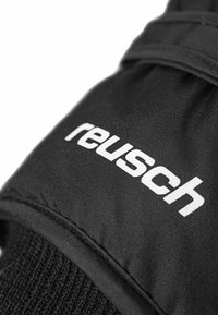 Svarta handskar med en texturerad yta och ribbat manschetter. Ordet "reusch" är tydligt visat i feta, vita bokstäver.