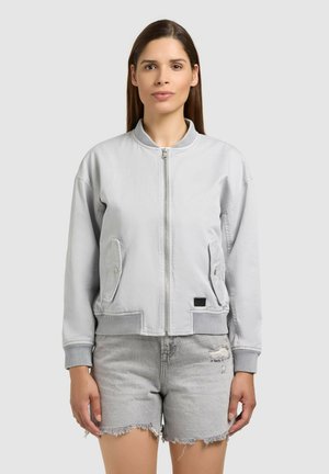 Nő áll előre nézve, világosszürke cipzáras bomber dzsekit és kopott szürke farmer rövidnadrágot visel egyszínű háttér előtt.