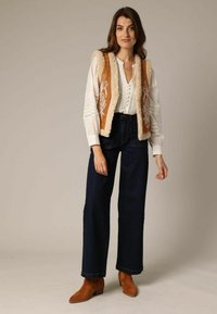 Gilet brodé marron avec bord crème porté sur une blouse blanche, associé à un jean large bleu foncé et des bottines marron.