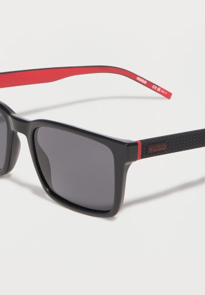Lunettes de soleil rectangulaires noires avec des verres gris et des branches intérieures rouges, présentant des branches extérieures noires texturées avec le logo rouge "HUGO".