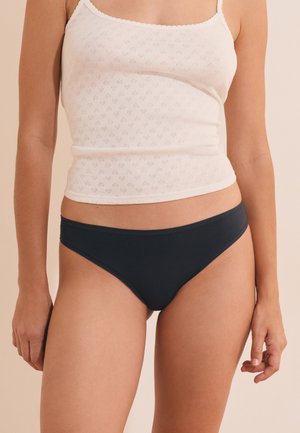 Vrouw draagt een wit tanktop met patroon en zwarte bikini-ondergoed, staand tegen een effen beige achtergrond.
