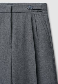Pantaloni grigi plissettati in un tessuto texturizzato, con chiusura a bottone e tasche laterali, che mostrano un design su misura e linee pulite.