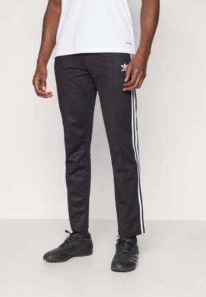 Mężczyzna ubrany w czarne spodnie dresowe Adidas z białymi paskami po bokach, czarne buty sportowe i białą koszulę na jednolitym tle.