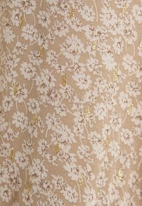 Beige stof met een delicate witte bloemmotief, geaccentueerd met gouden spikkels. De textuur lijkt zacht en geweven, met een subtiele glans.