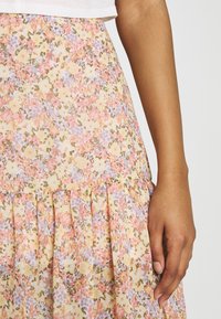 Jupe florale en tissu jaune avec des motifs roses, violets et verts. Présente des plis au niveau de l'ourlet et une taille ajustée. Texture lisse.