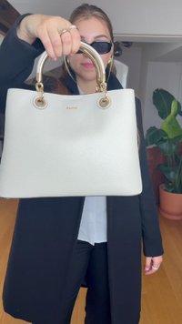 Kremowa skórzana torebka o strukturalnym kształcie, ze złotymi akcentami i okrągłymi uchwytami. Posiada logo z przodu oraz teksturowane wykończenie.