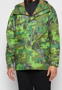 Green camo jacket featuring a mix of light and dark shades, zip-up front, multiple pockets, and textured fabric with a glossy finish.