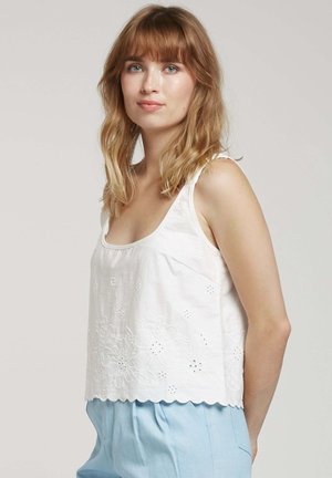 Haut sans manches blanc avec un ourlet festonné, présentant des détails de broderie florale. Fabriqué en tissu léger, associé à un pantalon bleu clair.