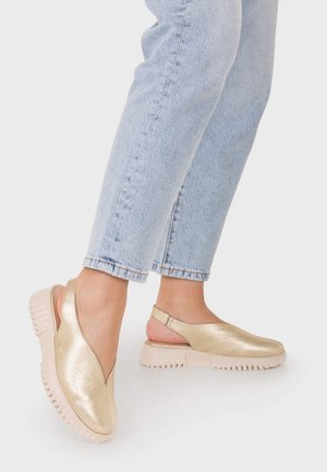 Pies con zapatos slingback dorados con suelas blancas gruesas y vaqueros cortos azul claro sobre un fondo liso.
