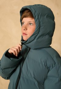 Junge mit Sommersprossen, der eine türkisfarbene Kapuzenjacke trägt, hält den Reißverschluss nahe seines Kinns und schaut nachdenklich zur Seite vor einem beigen Hintergrund.
