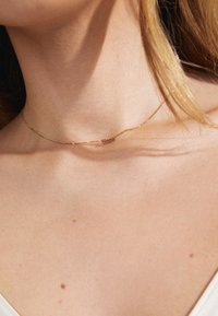 Zelta pārklājuma kaklarota ar smalku ķēdīti, uz kuras, rakstīts vārds "Cerība" kursīvā, atpūšas uz atklātas ādas tuvumā pie apkakles kaula.