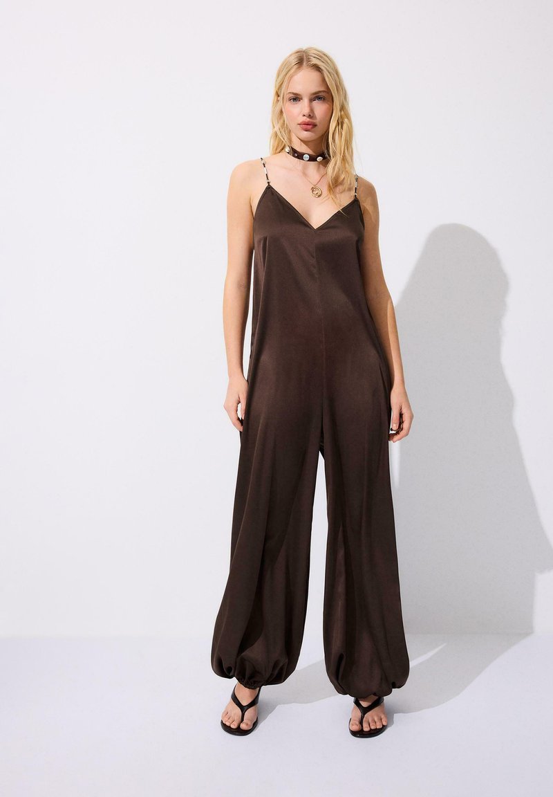 Femme blonde debout contre un mur blanc, portant une combinaison sans manches marron à coupe ample avec des ourlets froncés, des sandales noires, un collier ras du cou et un collier.
