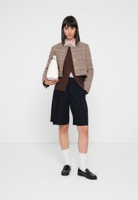 Kockás cropped dzseki, barna pulóver, csíkos ing, tengerészkék rövidnadrág, fekete papucs, fehér zokni, fehér táska hordása, sima háttéren.