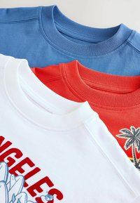 Trīs kokvilnas crewneck svārki: zili, sarkani un balti. Katrs ir ar ribotu apkakli un izteiktiem grafiskiem attēliem uz baltajiem un sarkaniem krekliem.