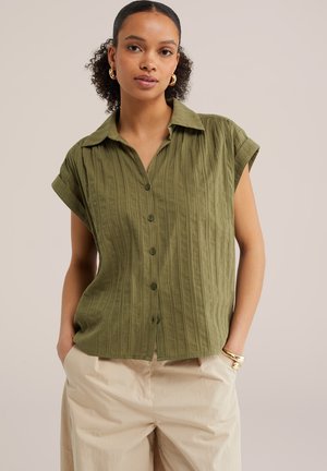 Žena s kučeravými vlasmi, ktorá nosí zlaté kruhové náušnice, olivovo zelenú krátkym rukávom zapínanú košeľu, béžové nohavice a zlaté náramky, stojí s rukami v kapsách.