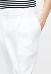 Γυναίκα που φοράει λευκό ψηλόμεσο πλισέ παντελόνι με πλαϊνές τσέπες και μαύρη-λευκή ριγέ μπλούζα, το χέρι της ακουμπά στην τσέπη.