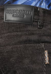 Detailný záber na hnedé kordové nohavice s čiernou koženou nášivkou Just Cavalli Milano a malou vzorovanou textilnou nášivkou Just Cavalli blízko vrecka.