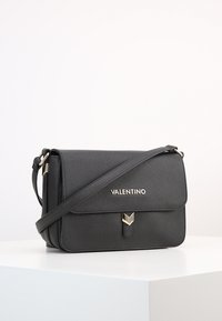 Svart läder crossbody-väska med en strukturerad design, detaljer i guld, främre klaff och logotypdetalj. Justerbar axelrem ingår.