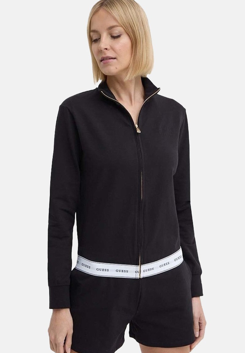 Chaqueta negra con cremallera dorada, con una cinturilla blanca con marca. Hecha de tejido suave, con mangas largas y un diseño ajustado.