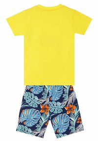 Camiseta amarilla de manga corta con cuello redondo, combinada con pantalones cortos marinos que presentan grandes hojas tropicales azules y verdes, y flores naranjas.